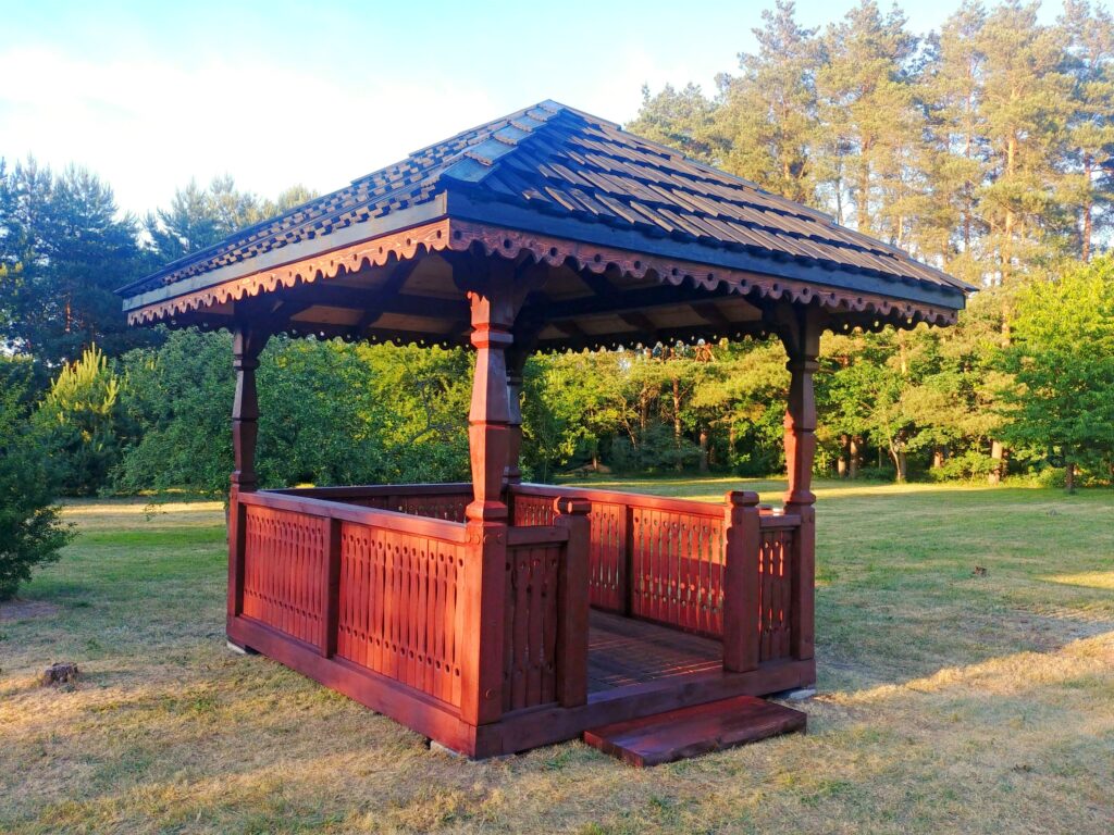 Drewniana altana z ozdobnym dachem, umiejscowiona na tle zielonego terenu z drzewami w tle.