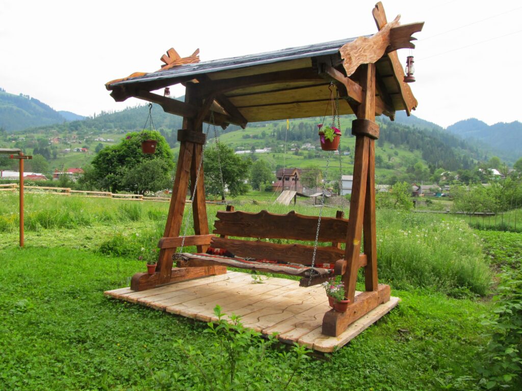 Drewniana huśtawka z dachem, zawieszona na jednej z platform w otoczeniu zielonych pól i gór, z wiszącymi doniczkami kwiatów.