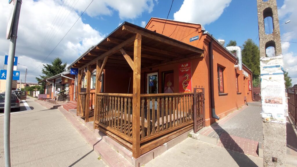 Zewnętrzny widok restauracji z pomarańczową elewacją i drewnianym zadaszeniem. Na podjeździe widoczna lada oraz reklama "Bar Dobrze Napięty Smak". Obok znajdują się znaki drogowe oraz zaparkowane samochody na chodniku, a w tle widać niebo z chmurami.