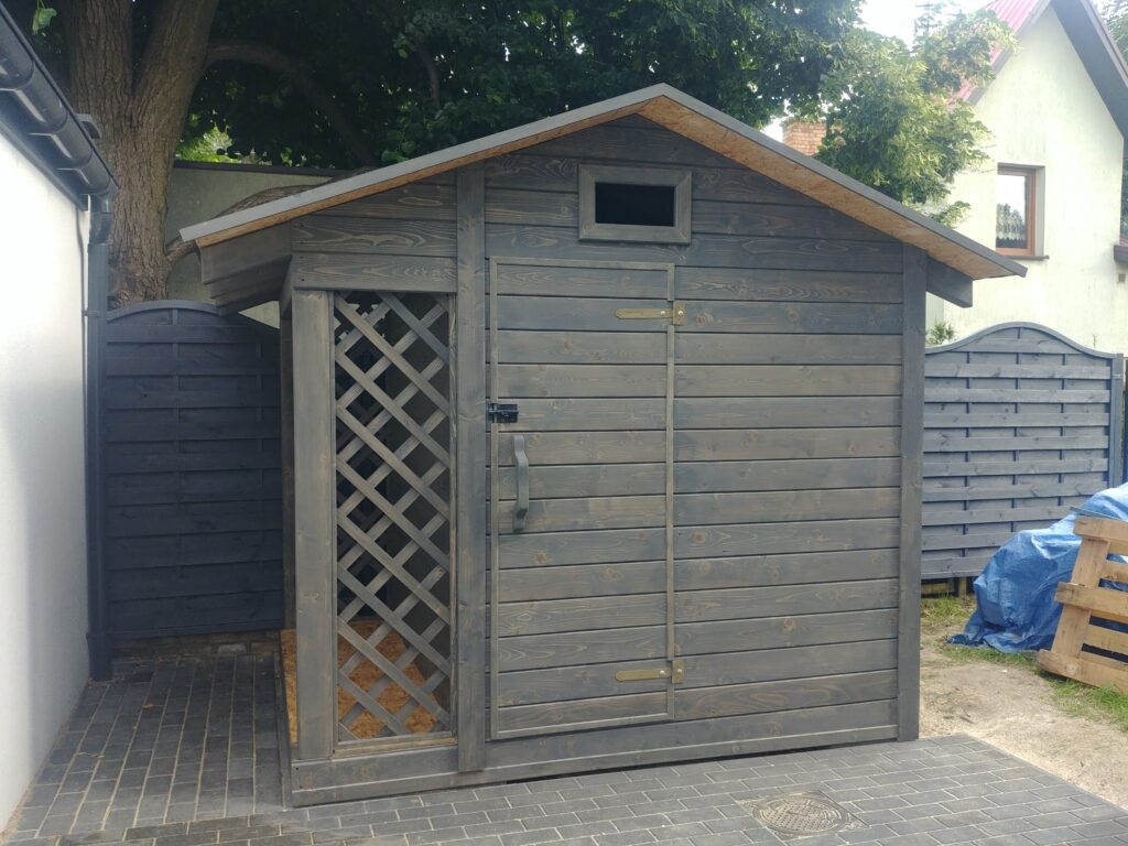 Szary drewniany domek ogrodowy z dachówką o spadzistym dachu, z drzwiami i kratownicą, ustawiony na kostce brukowej, w tle widoczne ogrodzenie i niebieska plandeka.
