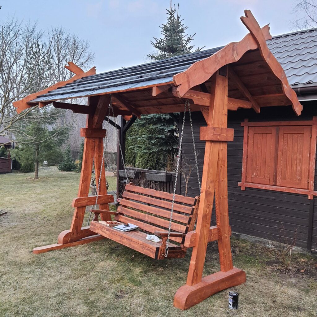Drewniana huśtawka z zadaszeniem, umiejscowiona w ogrodzie, na tle ciemnej chaty, z widocznymi drzewami w tle i trawnikiem.