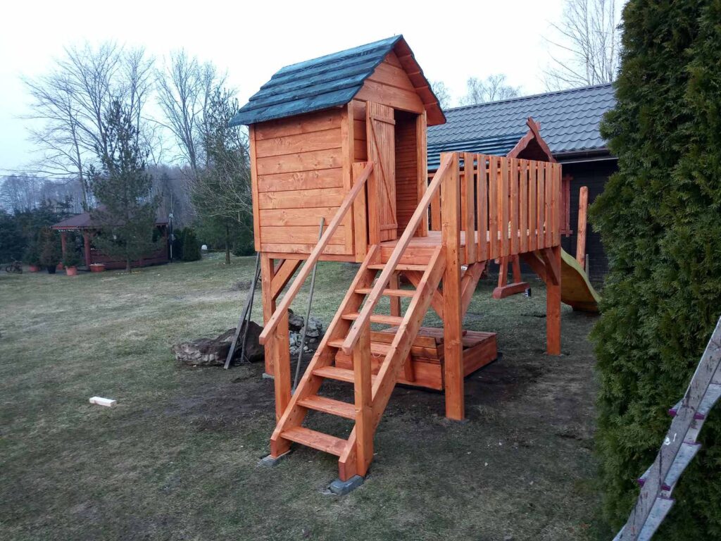 Drewniany plac zabaw z domkiem na wysokości, z drewnianymi schodami i zjeżdżalnią, otoczony trawą i drzewami, w tle widać drewnianą altankę.