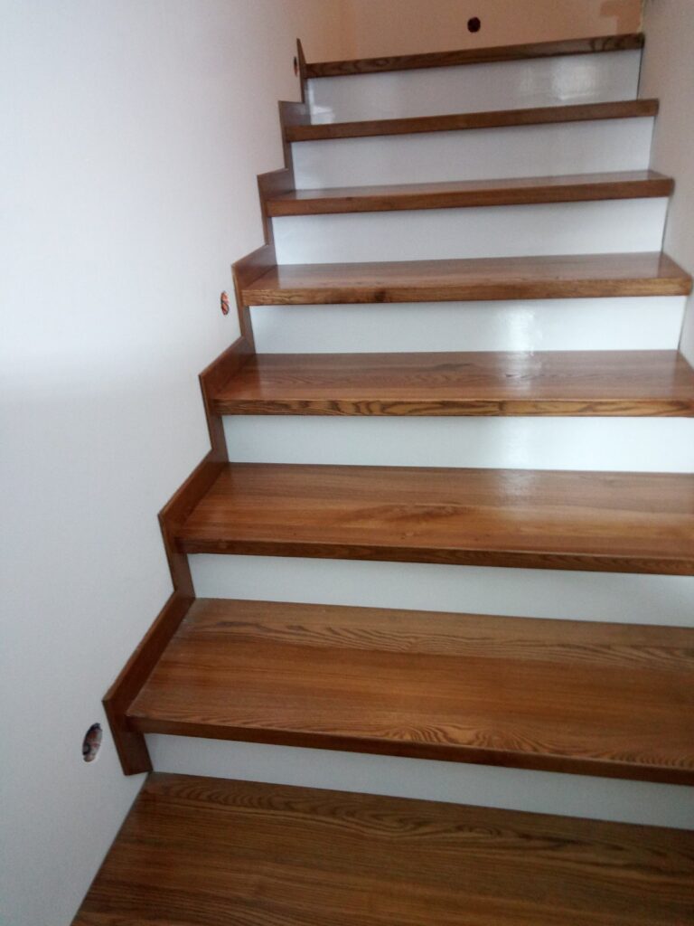 Widok drewnianych schodów prowadzących w górę, z jasno pomalowanymi bokami i widocznymi detalami drewna na stopniach, w tle jasna ściana.