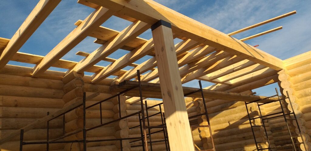 Widok wnętrza budowanego domku z bali, z widocznym drewnianym stropem i konstrukcją podtrzymującą; w tle niebieskie niebo z chmurami, a na dole stalowe rusztowanie.
