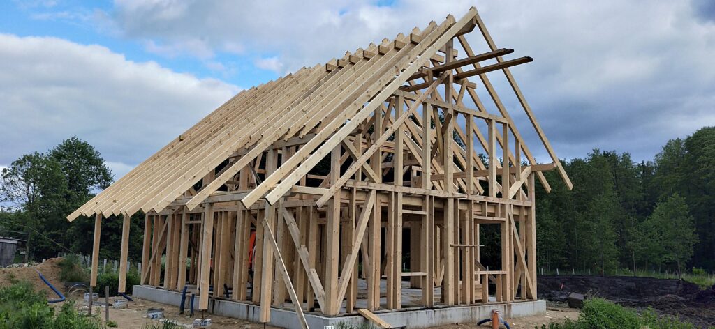 Niedokończony dom w konstrukcji drewnianej z widocznymi belkami i rozstawem, otoczony zielenią drzew i gruntami budowlanymi, pod niebieskim, lekko pochmurnym niebem.