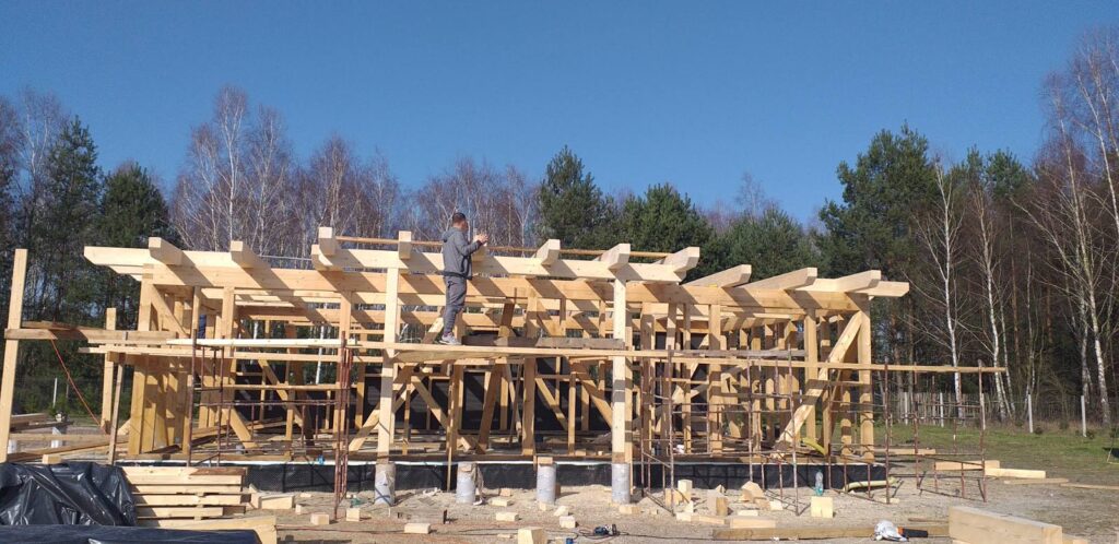 Robotnik na chwilę odpoczywa na dachu konstrukcji z drewnianych belek, otoczonej lasem i błękitnym niebem w tle. Widać budowę, na której użyto dużej ilości drewnianych materiałów oraz rusztowań.