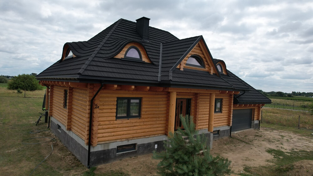Dwupoziomowy dom z bali drewnianych o ciemnym dachu z falistym kształtem, z oknami w kształcie łuków, znajdujący się w wiejskim otoczeniu z zielonym trawnikiem i drzewami w tle.