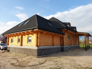 Widok z boku nowoczesnego domu z drewna, z charakterystycznymi oknami na dachu i pokryciem dachowym z czarnych dachówek, otoczonego piaskiem i zielonym trawnikiem w tle.