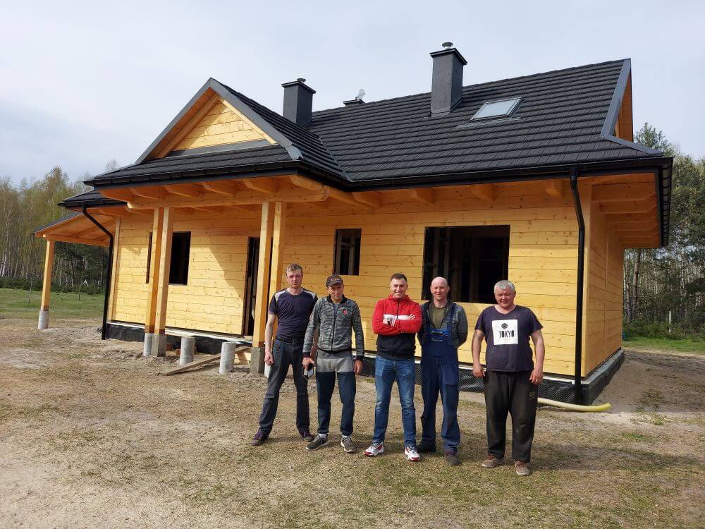 Grupa pięciu mężczyzn stoi przed nowym, drewnianym domem o czarnym dachu, otoczonym przez pole i drzewa w tle. Mężczyźni są ubrani w różne ubrania robocze, a niektórzy trzymają narzędzia. Dom ma duże okna i jest w fazie wykończeniowej.