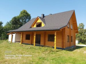 Nowy drewniany dom z czarnym dachem, w rozwijającym się otoczeniu zieleni, z widocznymi dużymi oknami i symbolami budowy z boku.