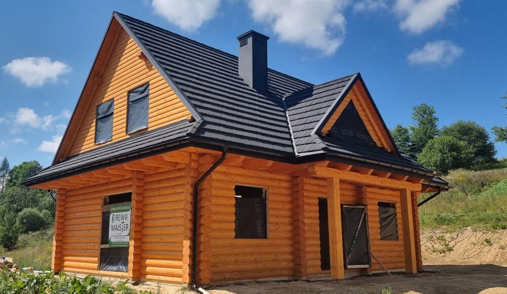 Nowy dom drewniany o podwójnym dachu, wykonany z bali, z czarnym dachem i widocznymi oknami, usytuowany w słonecznym otoczeniu z zielonymi drzewami w tle.
