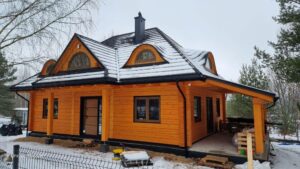 Widok na nowo wybudowany dom w stylu drewnianym, z pomarańczowym drewnem na elewacji i śniegiem na dachu; dom ma poddasze z oknami pod szczytem dachu oraz dużą zadaszoną werandę.