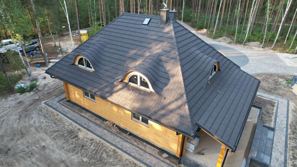 Widok z lotu ptaka na nowoczesny, jednopiętrowy dom z drewnianym obiciem i ciemnym, falistym dachem. Dach z małymi oknami dachowymi, otoczony lasem, z widocznymi ścieżkami i terenami budowy w pobliżu.