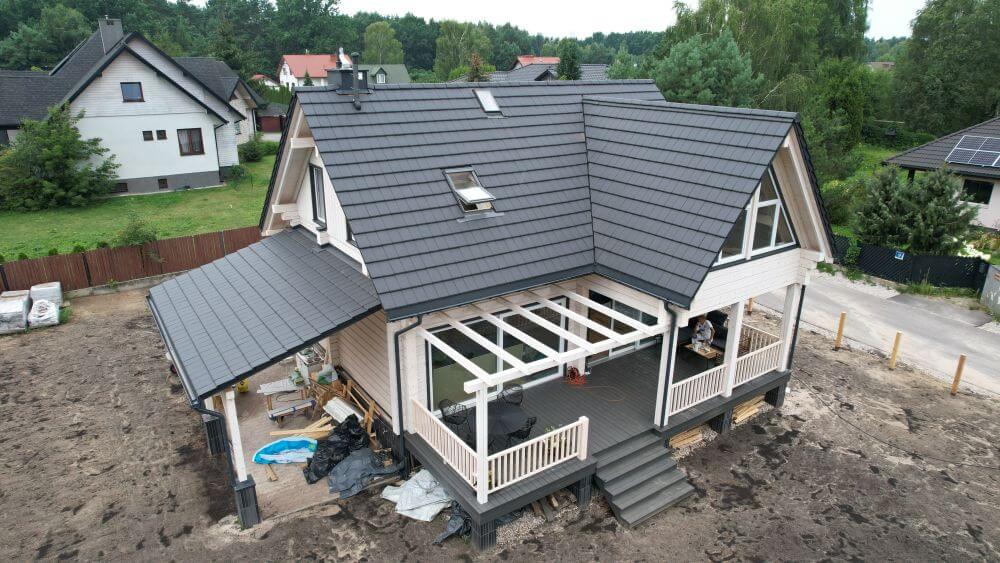 Widok na nowoczesny dom z czarnym dachem, tarasem z białymi balustradami i przyległym zadaszonym miejscem na świeżym powietrzu, otoczony gleba i inne budynki w tle.