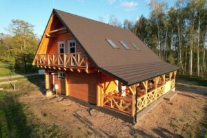 Widok na drewniany, dwu piętrowy dom z mansardowym dachem, z balkonem i dużymi oknami, otoczony zielenią i drzewami, z widocznymi meblami na tarasie.